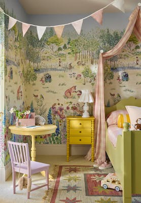 Children's bedroom featuring pastel mural wallpaper (Potter's Woodland - Spring) with a bed to the right and table and chair to the left with bunting hanging on the ceiling.